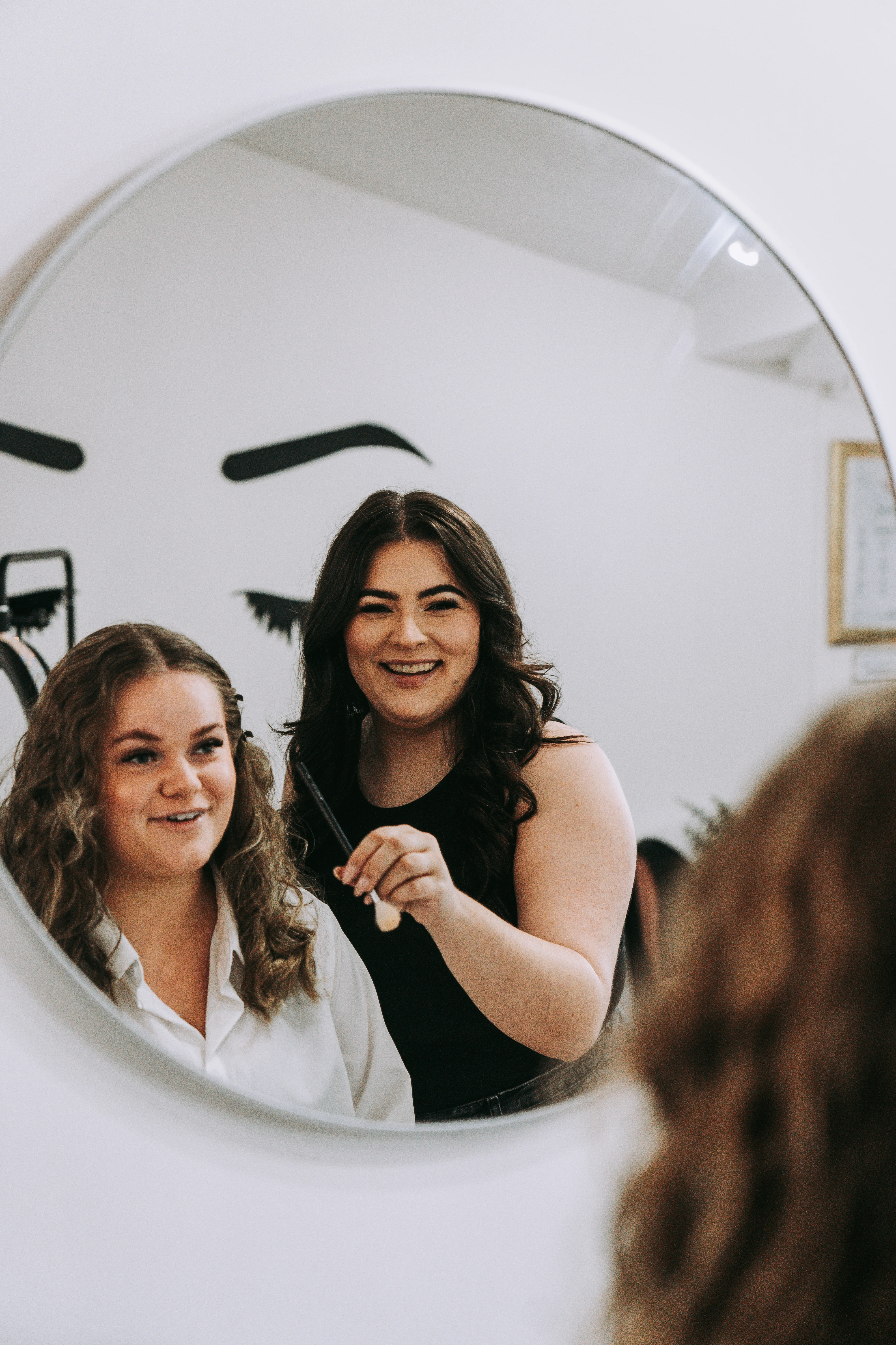 Jade and a client smiling, reflected in a round mirror at the Roselli Cosmetologist salon